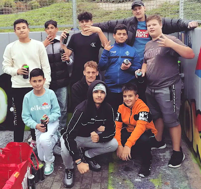 Die Jugendlichen, die die Wand hinter dem DFB-Fußballfeld mit Graffiti verschönert haben, gemeinsam mit dem Graffiti-Künstler Felix Wich (hinten) und Jugendsozialarbeiter Benjamin Reimer (Mitte). Graffitibande 2022